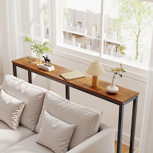 Könnte beinhalten: Ein langer, schmaler Konsolentisch mit einer dunkelbraunen Holzplatte und einem schwarzen Metallrahmen. Der Tisch steht vor einem Fenster und ist mit einer kleinen Lampe, einer Vase und anderen dekorativen Gegenständen dekoriert. Der Tisch ist 152 cm lang.