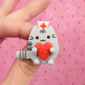 May include: A grey cat figurine wearing a nurse's hat and holding a red heart with a heartbeat symbol. The figurine is made of polymer clay.