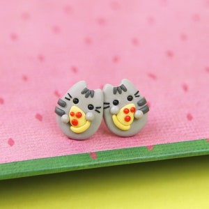 May include: A pair of gray cat-shaped earrings with a slice of pizza in their mouths. The pizza has red pepperoni and yellow cheese.