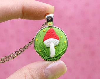 May include: A silver chain necklace with a green and red mushroom pendant. The mushroom has a white stem and red cap with white spots. The pendant is surrounded by green grass and yellow flowers.