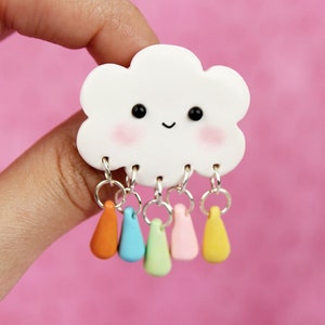 Könnte beinhalten: Eine weiße Wolke mit einem lächelnden Gesicht und rosafarbenen Wangen, an der fünf bunte Regentropfen hängen. Die Wolke ist aus Polymer Clay gefertigt und die Regentropfen sind aus Polymer Clay in Orange, Blau, Grün, Rosa und Gelb.