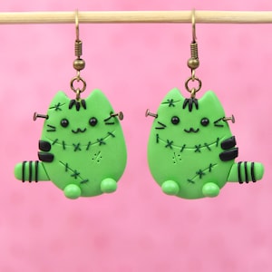 May include: A pair of green cat-shaped earrings with black stitching and black and white stripes. The earrings are hanging from gold hooks.