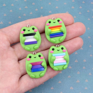 May include: Four green frog charms with different pride flags on their backs. The flags are the transgender flag, the bisexual flag, the pansexual flag, and the rainbow pride flag.