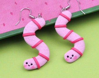 May include: A pair of pink and red worm earrings with a cute face. The earrings are made of polymer clay and have a silver hook.