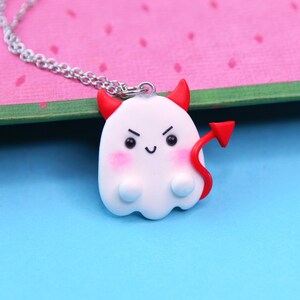 May include: A white ghost pendant necklace with red devil horns and a red tail. The pendant features a small, smiling face with black eyes and pink cheeks. The necklace chain is silver.