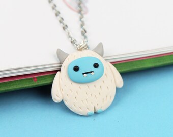 May include: A silver chain necklace with a white and blue yeti charm. The yeti has a blue face with a small smile and two small black eyes.