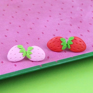 May include: Two pairs of polymer clay earrings shaped like strawberries. One pair is pink and the other is red.
