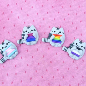 May include: Four gray cat figurines holding different pride flags. The flags are a rainbow flag, a transgender flag, a bisexual flag, and a pansexual flag.