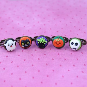 May include: Five Halloween-themed rings with bronze-colored adjustable bands. The rings feature a ghost, black cat, cauldron, pumpkin, and skull. Each ring has a round, colorful design. The background is a pink surface with small white dots.