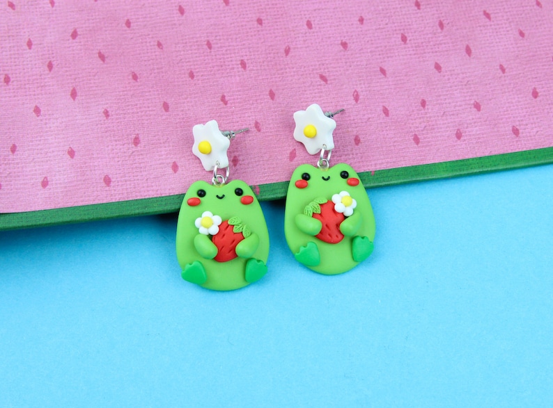 May include: A pair of green frog earrings with white flower accents. Each frog is holding a red strawberry.