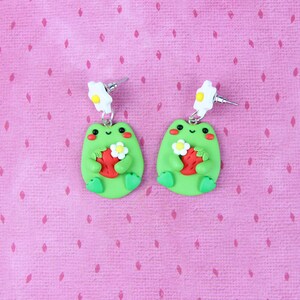 May include: A pair of green frog earrings with white flower accents. Each frog is holding a red strawberry with green leaves. The earrings are on a pink background.