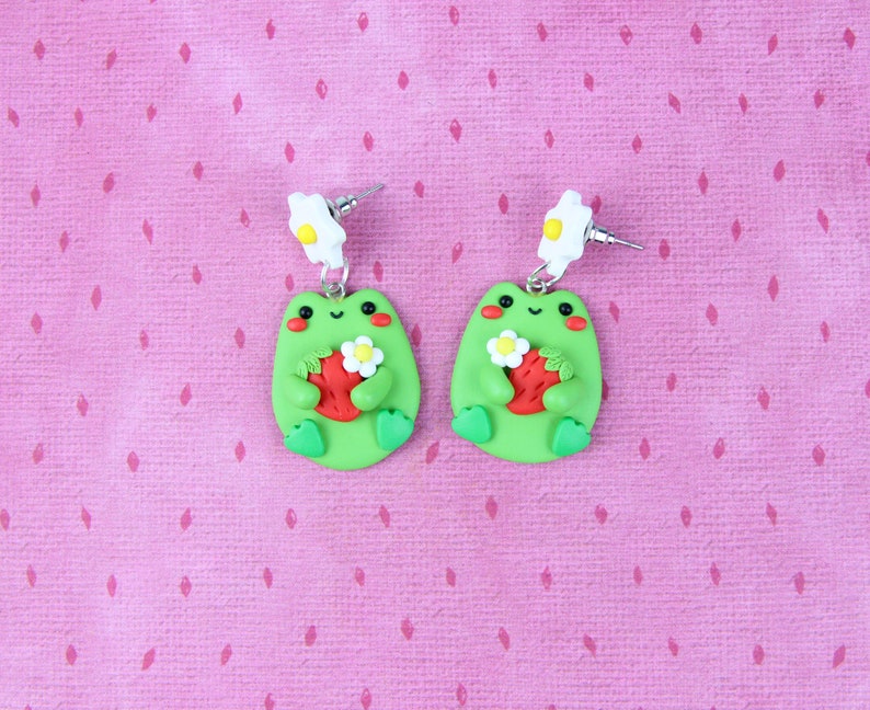 May include: A pair of green frog earrings with white flower accents. Each frog is holding a red strawberry with green leaves. The earrings are on a pink background.