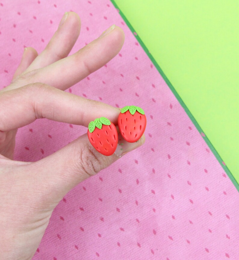 May include: A pair of red and green strawberry-shaped earrings. The earrings are made of polymer clay and are about 1 inch in diameter.