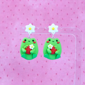 May include: A pair of green frog earrings with white flowers and red strawberries. The frogs are holding the strawberries in their hands.