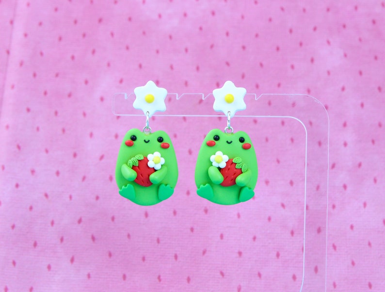 May include: A pair of green frog earrings with white flowers and red strawberries. The frogs are holding the strawberries in their hands.