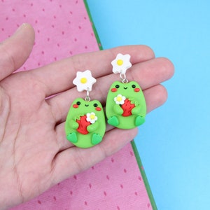 May include: A pair of green frog earrings with white flower accents. Each frog is holding a red strawberry.