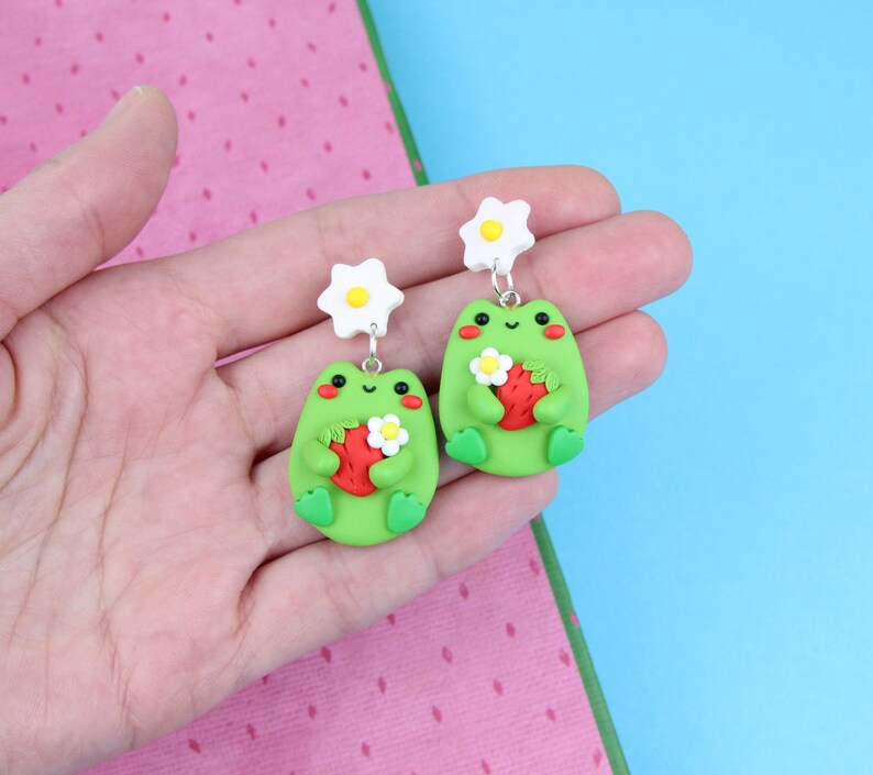 May include: A pair of green frog earrings with white flower accents. Each frog is holding a red strawberry.