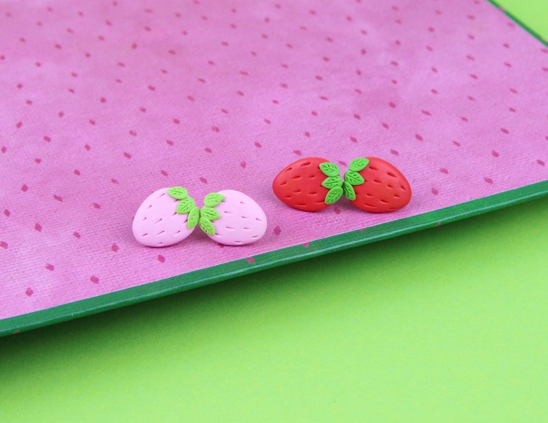 May include: Two pairs of polymer clay earrings shaped like strawberries. One pair is pink and the other is red.