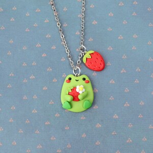 May include: A green frog charm necklace with a silver chain. The frog is holding a red strawberry and a white flower. The necklace also has a red strawberry charm.