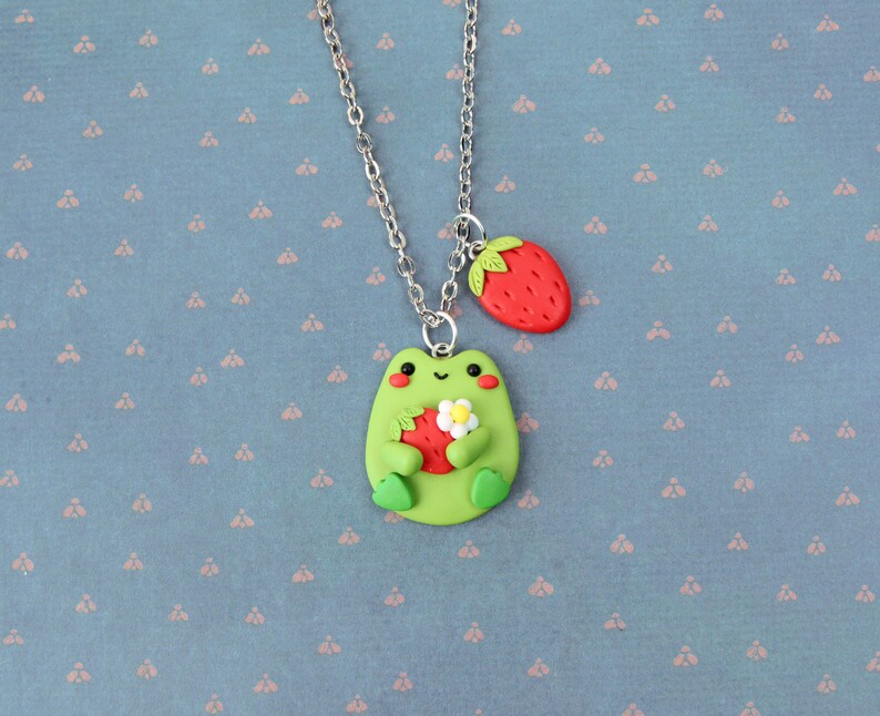 May include: A green frog charm necklace with a silver chain. The frog is holding a red strawberry and a white flower. The necklace also has a red strawberry charm.