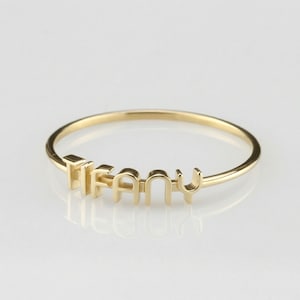 May include: A gold-colored ring with the name "TIFFANY" spelled out in individual capital letters. The ring is thin and delicate, with the letters slightly raised above the band. The ring is set against a white background.