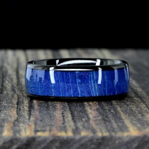 Könnte beinhalten: Ein schwarzer Wolframcarbid-Ring mit einer blauen Holzeinlage. Der Ring hat eine polierte Oberfläche und eine abgeschrägte Kante.