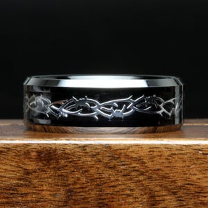 Könnte beinhalten: Ein schwarzer Ring mit einem silbernen Dornendesign. Der Ring hat eine polierte Oberfläche und liegt auf einer Holzoberfläche. Das Dornendesign umschließt das gesamte Band.