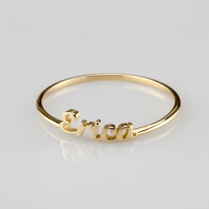 May include: A delicate gold ring featuring the name "Erica" in a cursive font. The ring is a thin band with the name as a prominent design element, showcasing a personalized jewelry piece.