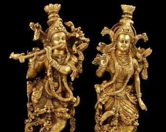May include: A pair of gold-toned metal statues of Hindu deities Radha and Krishna. Krishna is playing a flute and Radha is holding a flower. The statues are standing on ornate bases.
