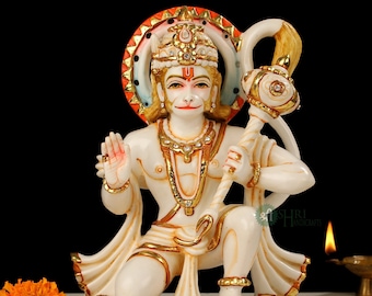 Może przedstawiać: Biały marmurowy posąg hinduskiego bóstwa Hanuman, trzymający złotą laskę z ozdobnym, wysadzanym klejnotami wierzchołkiem. Ma na sobie czerwoną i złotą koronę oraz białą i złotą szatę. Posąg stoi na białym marmurowym cokole z złotymi akcentami.