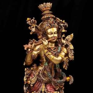 May include: A gold-colored statue of Krishna playing a flute, adorned with intricate details and colorful gemstones. The statue is set against a black background.