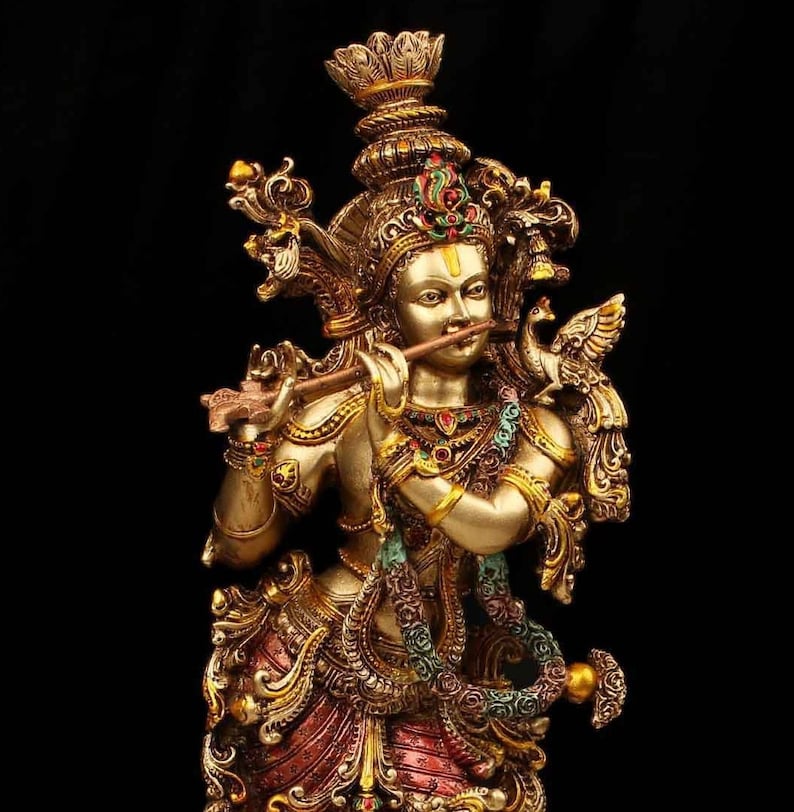 May include: A gold-colored statue of Krishna playing a flute, adorned with intricate details and colorful gemstones. The statue is set against a black background.