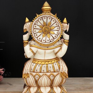 May include: White marble statue of the Hindu goddess Lakshmi, sitting on a lotus base. She is adorned with gold accents and a circular halo with a floral design.