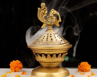 May include: A gold metal incense burner with a peacock design on top. The burner is emitting smoke and is surrounded by orange marigold flowers on a white surface. The text "Shree Hari Handicrafts" is visible on the burner.