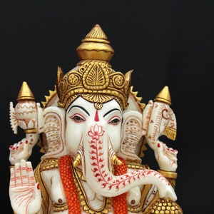 May include: White marble statue of the Hindu deity Ganesha, with a gold crown and red and white accents. The statue is intricately carved and features a trunk, large ears, and a single tusk.