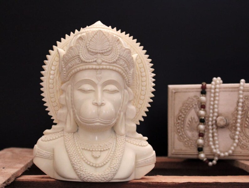 May include: White ivory bust sculpture of the Hindu deity Hanuman, with a crown and ornate jewelry. The sculpture is on a wooden surface.