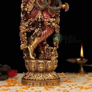 May include: A gold-toned statue of a Hindu deity, possibly Krishna, with intricate details and a multi-colored base. The statue is standing on a lotus-shaped base and is adorned with jewels and flowers.