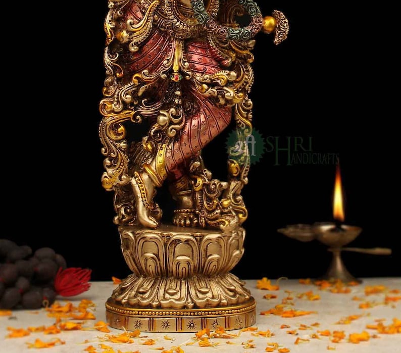 May include: A gold-toned statue of a Hindu deity, possibly Krishna, with intricate details and a multi-colored base. The statue is standing on a lotus-shaped base and is adorned with jewels and flowers.