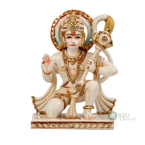 Może przedstawiać: Biały marmurowy posąg hinduskiego b&oacute;stwa Hanuman, klęczącego z uniesioną prawą ręką w geście błogosławieństwa. Trzyma w lewej ręce złotą laskę. Posąg jest ozdobiony złotą farbą i kryształkami. Podstawa posągu jest z czerwonego i białego marmuru z pozłacanymi zdobieniami.