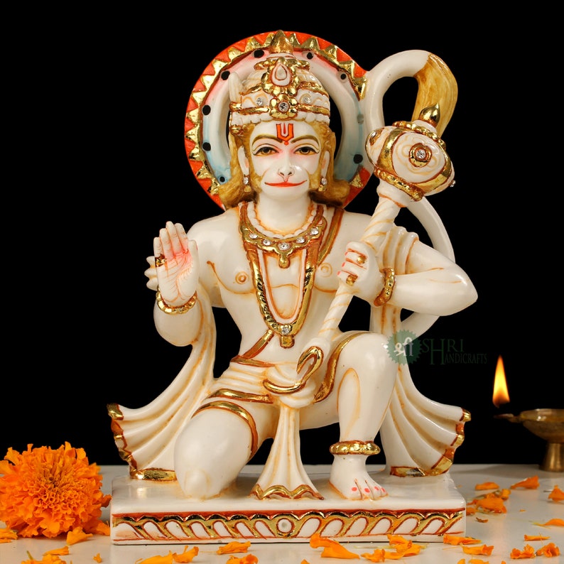 Może przedstawiać: Biała marmurowa statua hinduskiego b&oacute;stwa Hanuman, klęczącego z złotą laską w prawej ręce i uniesioną prawą ręką w geście błogosławieństwa. Nosi złotą koronę i naszyjnik z kamieniami szlachetnymi. Statua stoi na białej podstawie z czerwoną i złotą obw&oacute;dką.