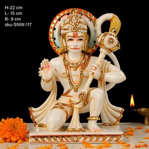 Może przedstawiać: Biały marmurowy posąg Hanuman, hinduskiego b&oacute;stwa, siedzącego z uniesioną prawą ręką w geście błogosławieństwa. Trzyma maczugę w lewej ręce. Posąg jest ozdobiony złotą farbą i kryształkami. Posąg ma 22 cm wysokości, 15 cm szerokości i 9 cm głębokości. sku-SNW-117