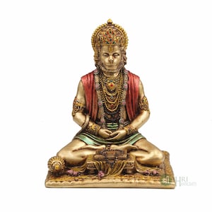 Może przedstawiać: Złota statua hinduskiego boga Hanuman, siedzącego w medytacyjnej pozie. Ma na sobie czerwoną i zieloną szatę, koronę i wiele naszyjnik&oacute;w. Siedzi na złotej platformie z książką i małą kulą.