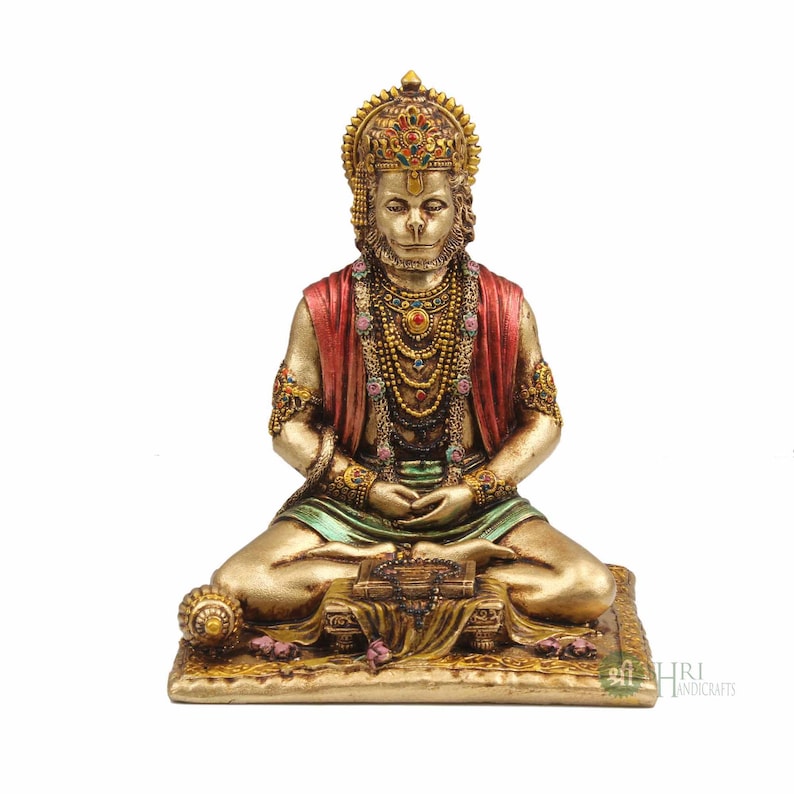 Może przedstawiać: Złota statua hinduskiego boga Hanuman, siedzącego w medytacyjnej pozie. Ma na sobie czerwoną i zieloną szatę, koronę i wiele naszyjnik&oacute;w. Siedzi na złotej platformie z książką i małą kulą.