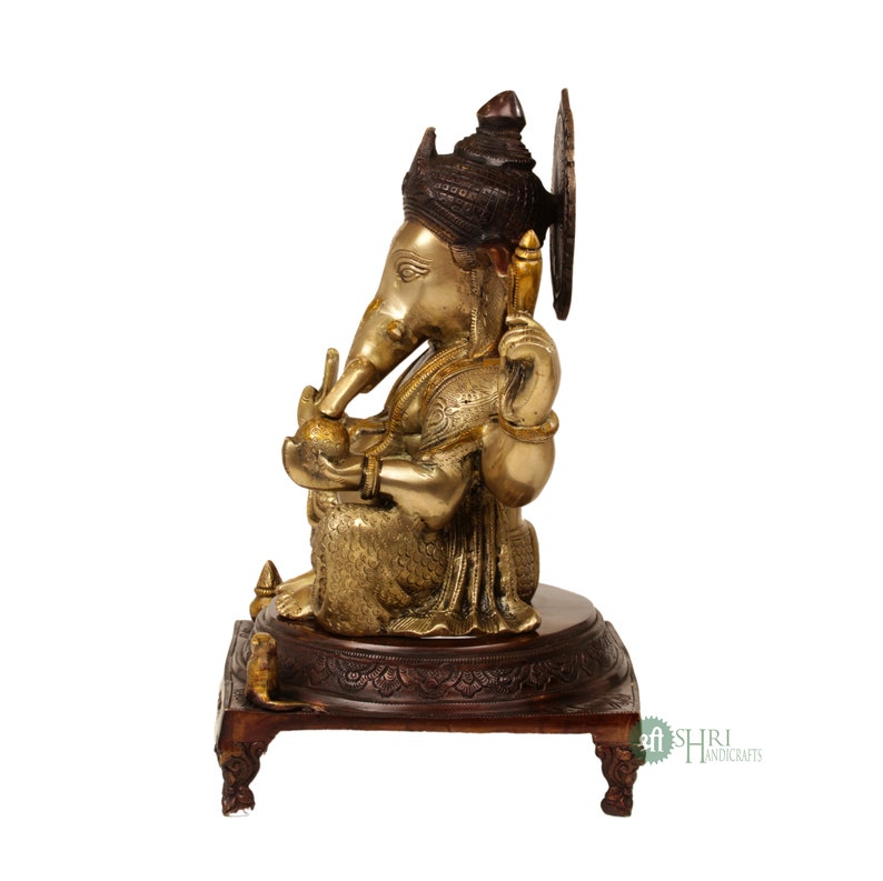 May include: A gold-toned brass statue of Ganesha, the Hindu god of wisdom and prosperity, sitting on a wooden base. The statue is intricately detailed and features a crown, a trunk, and a broken tusk.