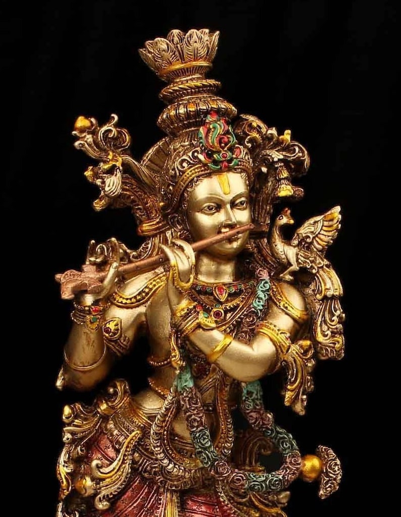 May include: A gold-toned statue of Krishna playing a flute. The statue is intricately detailed with colorful accents and a peacock feather headdress.
