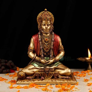 Może przedstawiać: Złota statua hinduskiego boga Hanuman, siedzącego w medytacyjnej pozie. Ma na sobie czerwoną i zieloną szatę, koronę i wiele naszyjnik&oacute;w. Statua stoi na złotej podstawie z kwiatowym wzorem.