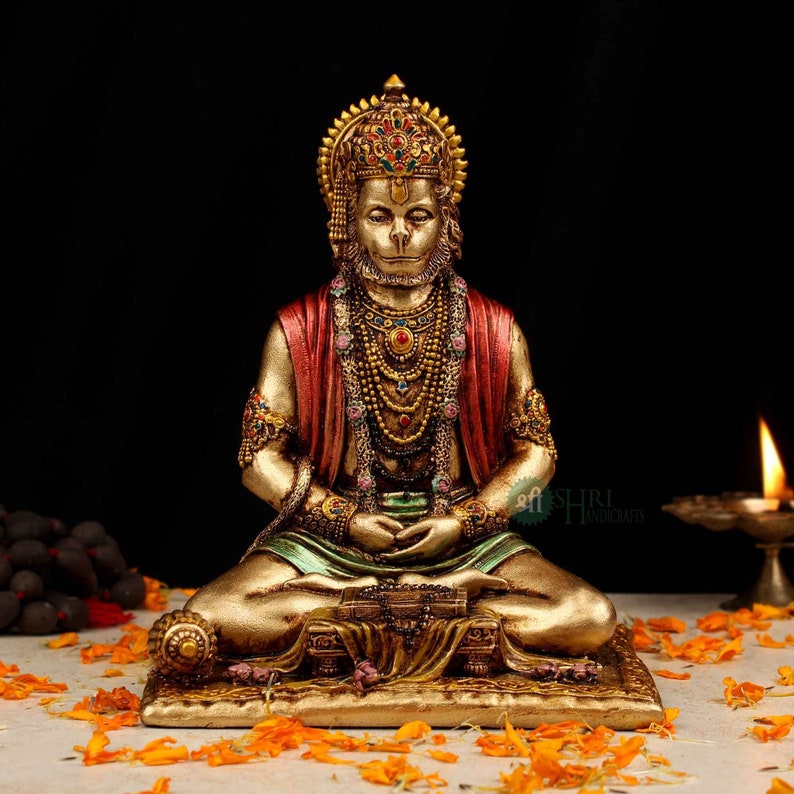 Może przedstawiać: Złota statua hinduskiego boga Hanuman, siedzącego w medytacyjnej pozie. Ma na sobie czerwoną i zieloną szatę, koronę i wiele naszyjnik&oacute;w. Statua stoi na złotej podstawie z kwiatowym wzorem.