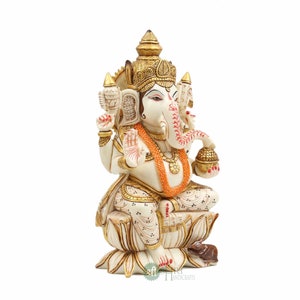 May include: A white and gold Ganesha statue sitting on a lotus flower. The statue is decorated with intricate details and has a red and gold garland around its neck. The statue is sitting on a white base with a small mouse at its feet.