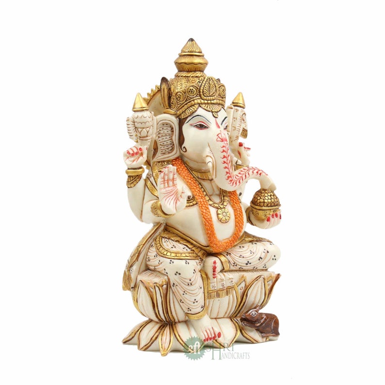May include: A white and gold Ganesha statue sitting on a lotus flower. The statue is decorated with intricate details and has a red and gold garland around its neck. The statue is sitting on a white base with a small mouse at its feet.