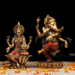 Puede incluir: Estatua dorada de Lakshmi, la diosa hindú de la riqueza, sentada sobre una flor de loto, sosteniendo una olla de oro.  Está junto a una estatua dorada de Ganesha, el dios hindú de los comienzos, bailando sobre una flor de loto. Las estatuas están sobre una superficie blanca con pétalos de flores naranjas.